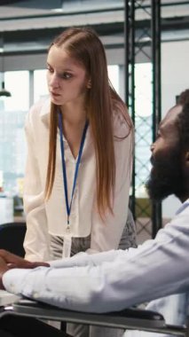 Dikey video Junior Dev ve tekerlekli sandalyede çalışan iş arkadaşı yazılım uygulamaları geliştiriyor, kodu optimize ediyor. Belden aşağısı felçli kadın teknisyen ve meslektaşı yapay zeka kullanarak dar boğazları ve kameraları çözüyor.