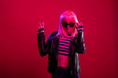 Deri ceketli ve güneş gözlüklü kendine güvenen çekici kadın stüdyoda kaygısız punk enerjisiyle modellik yapıyor. Siyah gölgesine dokunan ve barış işareti el hareketleriyle poz veren acayip bir kadın manken..