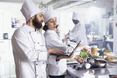 Yeni yemek tariflerini dizüstü bilgisayarda araştıran şapkalı uzmanlardan oluşan bir ekip malzemeleri hazırlamadan, taze ürünleri doğramadan ve yeni gurme yemekleri yaratmadan önce. Lokanta mutfağında aşçılık sanatı.