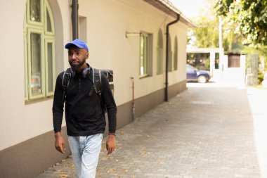 Termal sırt çantalı Afrikalı Amerikan kuryesi yemek siparişi vermek için kapıya yaklaşıyor. Akıllı telefon uygulaması kullanarak teslimat yerini doğruladı ve dahili telefonu çaldı..
