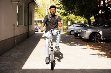 Sırt çantasında fast food siparişi getirmek için bisiklete binen siyah teslimatçı müşteriye yemek getirmek için teslimat bisikletini kullanıyor. Şehir genelinde hızlı servis sağlanıyor, kurye çantası..