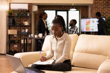 Siyahi kadın çalışan, bir iş planı hazırlamak için dijital aletleri ve evrakları birleştirerek panodan doküman okuyor. Afro-Amerikalı iş kadını ofis koltuğunda oturmuş basılı raporları inceliyor..