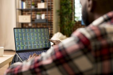 Uzaktan kumandalı tüccar oturma odasında oturmuş dizüstü bilgisayardaki hisse senedi indekslerini ve kripto para birimini takip ediyor. Evden çalışan Afrikalı Amerikalı adam, ticaret piyasası eğilimlerini ve yatırım stratejilerini inceliyor.