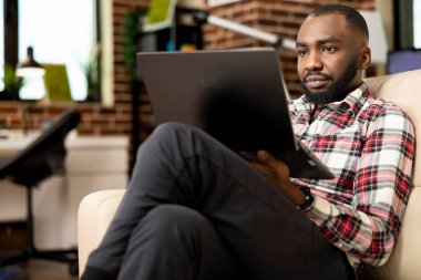 Afrika kökenli Amerikalı iş adamı bacak bacak bacak üstüne atmış, evden çalışıyor, başlangıç projesi raporları hazırlıyor. Odaklanmış serbest çalışan dizüstü bilgisayarını koltukta kullanır, özgeçmişi günceller ve iş olanaklarını tarar.