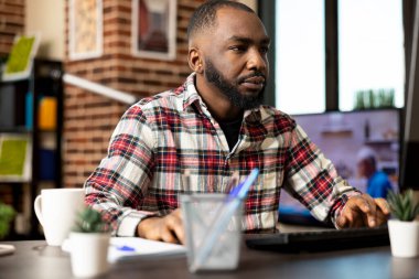 Evden çalışan odaklanmış siyahi bir adam, şirket kayıtlarını masaüstü bilgisayarında inceliyor. Elleri klavye ve farede olan Afrikalı Amerikalı girişimci, iş mali raporlarını son teslim tarihinden önce güncelliyor.
