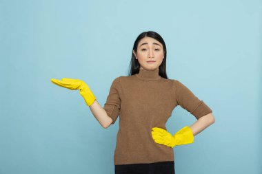 Sarı lastik eldivenli Asyalı temizlikçi bir eli belinde, diğeri de solda poz veriyor. Kadın temizleyici tanıtım içeriği için mavi arkaplan üzerinde izole kopya alanı sunuyor.