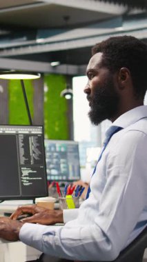 Açılış ofisinde yapay zeka dili model yardımı ile düşey video mühendisliği kodlaması, müzik dinlemek. Kulaklık takan Afrikalı Amerikalı adam LLM asistanı önerilerini kod düzenleyicisine entegre ediyor