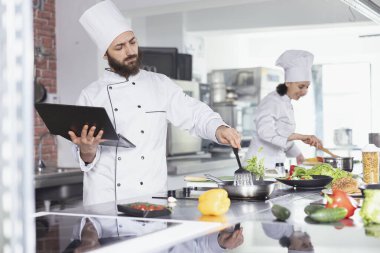 Modern bir mutfakta profesyonel aşçı geleneksel aşçılığı dijital aletlerle birleştiriyor, yeni tarifler keşfetmek için dizüstü bilgisayar kullanarak taze malzemeler hazırlıyor. Fırında pişirme uzmanı..