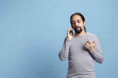 Orta Doğulu bir adam, bir arkadaşıyla telefonda konuşurken mavi arka planda duruyor, stüdyoda telefon hattında eğleniyor. Uzun saçlı sıradan bir yetişkin, insanları arıyor..