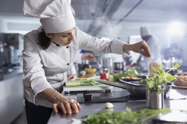 Modern bir restoranın mutfağında, önlüklü kadın aşçı, kaplanmış bir tabağı bitki yapraklarıyla süslüyor. Renkli sebzeler, mutfak aletleri ve taze ürünlerle dolu paslanmaz çelik çalışma alanı..