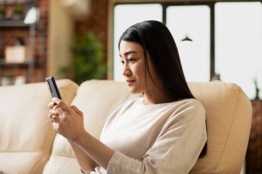 Asyalı girişimci kadın evdeki koltukta oturuyor, cep telefonunu tutuyor ve sosyal medyayı kontrol ediyor. Uzaktan çalışan bayan çalışanın ev işi sırasında dizüstü bilgisayarına ara vermesi.