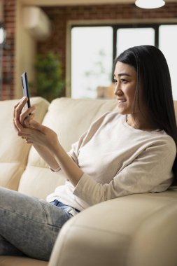 Asyalı bayan girişimci elinde akıllı telefonuyla rahat bir dairede oturmuş, video görüşmesi yapan yatırımcılarla proje fikirlerini tartışıyor. Sanal iş toplantısına katılan gülümseyen uzaktan kumandalı işçi.