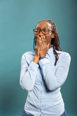 Mavi gömlek ve gözlük takan yorgun siyah bir kadın. Ağzını eliyle kapatıyor ve izole edilmiş arka planda esniyor. Gözleri kapalı, uyanık kalmaya çalışan Afrikalı Amerikalı kadın..