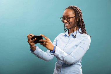 Mavi gömlekli gülümseyen genç kadın, izole edilmiş arka planda bir video izleyerek akıllı telefonuna derinden odaklanmış durumda. Neşeli Afrikalı Amerikalı bayan stüdyoda interaktif mobil oyunlar oynuyor..
