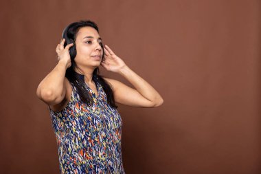 Hintli kadın birey yan bakıyor, kulaklıklarına dokunuyor ve dikkatle podcast dinliyor. Kablosuz kulaklık kullanan odaklanmış bir kadın. Stüdyoda kahverengi arka planda tek başına duruyor..