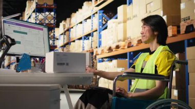 Tekerlekli sandalyedeki Asyalı adam talimatları işleme koyuyor ve kutuların dağıtım merkezindeki dağıtım için hazırlanıyor. Ticaret lojistik tesisinde yelek giyiyor. Hızlı teslimat. Kamera A.