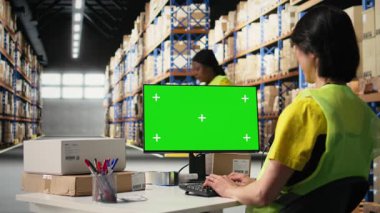 Asyalı personel paket izleme yazılımındaki izole ekran nakliye rotasına bakıyor. Hava yolu faturalarındaki teslimat detaylarını güncelliyor. Depoda çalışıyor, bilgisayarında yeşil ekran var. Kamera A.