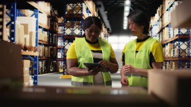 Afro-Amerikan ve Asyalı ekip sevkiyatı dijital envanter ve kargo paletleriyle birlikte depo raflarına hazırlayın. Ekspres teslimat için büyük ölçekli bir dağıtım merkezinde çalışıyor. Kamera A.