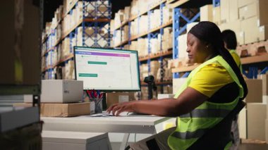 Afro-amerikan çalışan sipariş lojistik yazılımını güncelliyor, sevkiyat etiketlerini dağıtım için hazırlıyor. Ticaret lojistik tesisinde çelik yeleği olan endüstriyel depo personeli. Kamera A.