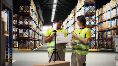 Çok ırklı çalışanlar malları kutuların içinde bir videoda tanıtıyorlar, depodaki endüstriyel depolama alanlarının yakınında canlı yayın yapıyorlar. Büyük bir iş tanıtımı reklamı. Kamera A.