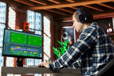 Kulaklık takan, video montajı yapan, evden yeşil ekran bilgisayarda çalışan bağımsız bir video editörü. Adam film projesinin görüntülerini düzenlemek için ses kulaklığı kullanıyor.