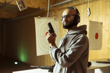 Arap atış poligonu müşterisi ateşli silah tutarak ev içi atış deneyiminin tadını çıkarıyor. Orta Doğulu adam koruyucu giysiler giyip güvenli ve hoş geldin ortamında silahlarla pratik yapıyor.