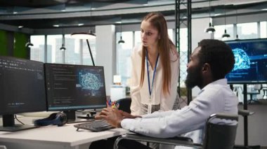 Küçük Dev ve tekerlekli sandalyedeki iş arkadaşı yazılım uygulamaları geliştiriyor, kodu optimize ediyor. Belden aşağısı felçli kadın teknisyen ve meslektaş, A kamerasındaki darboğazları çözmek için yapay zeka kullanıyor.