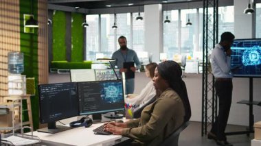 Başlangıç ofisindeki bilgisayar bilimci yapay zeka sistem ölçümlerini gözden geçiriyor, performans gösterge panellerini izliyor. Afrika kökenli Amerikalı bir kadın, bilgisayar ve kamera A 'daki araçları kullanarak sinirsel ağları ayarlıyor.