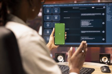 Masada oturan kadın, modern şehir merkezindeki AI LLLM sohbet robotu yazılımı ve yeşil ekran telefon kullanıyor. Apartmanda bilgisayar üzerinde büyük dil model programı ve akıllı telefon kullanılıyor