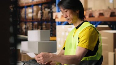 Yelek giyen bir adam endüstriyel depodaki sevkiyat etiketlerinin detaylarını inceler. Depodaki paketlerden gelen paketler ve pürüzsüz ekspres teslimat için takip numaraları. Kamera B.