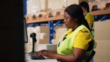 Afro-amerikan çalışan sipariş lojistik yazılımını güncelliyor, sevkiyat etiketlerini dağıtım için hazırlıyor. Ticaret lojistik tesisinde çelik yeleği olan endüstriyel depo personeli. Kamera B.