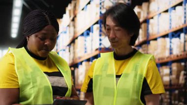 Afro-Amerikan ve Asyalı ekip sevkiyatı dijital envanter ve kargo paletleriyle birlikte depo raflarına hazırlayın. Ekspres teslimat için büyük ölçekli bir dağıtım merkezinde çalışıyor. Kamera B.