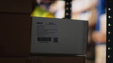 Tatmin merkezi personeli sipariş takibi için havayolu faturalarını tarıyor, depodaki nakliye etiketlerinde tarayıcı kullanıyor. Depoda raflı yelek giyen bir işçi. Kamera B.