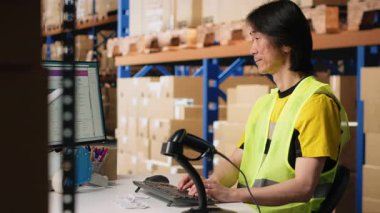 Asyalı bir çalışan, sanayi deposunda hava yolu faturalarını kaydediyor, paket izleme yazılımının sevkiyat detayları için barkod taraması yapıyor. Tatmin merkezi envanter takibi. Kamera B.