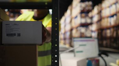 Tatmin merkezi personeli sipariş takibi için havayolu faturalarını tarıyor, depodaki nakliye etiketlerinde tarayıcı kullanıyor. Depoda raflı yelek giyen bir işçi. Kamera A.