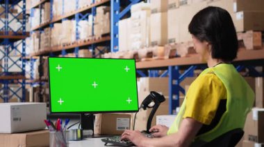 Asyalı personel paket izleme yazılımındaki izole ekran nakliye rotasına bakıyor. Hava yolu faturalarındaki teslimat detaylarını güncelliyor. Depoda çalışıyor, bilgisayarında yeşil ekran var. Kamera B.