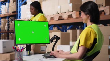 Personel veri girişi, envanter izleme motifleri ve görsel veya barkod yazıcı arayüzleri için depo odasındaki krom anahtar monitörünü gözden geçirir. Lojistik işçisi kargoyu ön planda paketler. Kamera B.