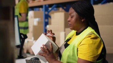 Afro-Amerikalı bir kadın kutulara havayolu faturası basıyor ve uyguluyor, rafların yakınındaki bir depoda yelek giyiyor. Sipariş yerine getirmek için takip bilgileriyle barkod etiketleri yönetiliyor. Kamera B.