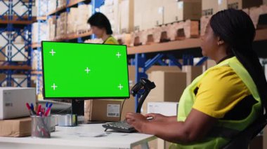 Siyah kadın işçiler yeşil ekran, depo otomasyon araçları ve envanter kontrol panellerinin yanında sipariş veriyor. Depolama raflarıyla birlikte e-ticaret emri işlemleri. Kamera B.