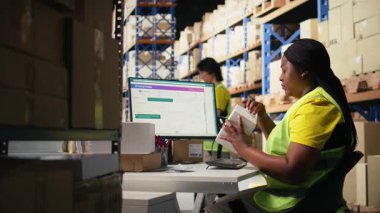 Afro-Amerikalı bir kadın kutulara havayolu faturası basıyor ve uyguluyor, rafların yakınındaki bir depoda yelek giyiyor. Sipariş yerine getirmek için takip bilgileriyle barkod etiketleri yönetiliyor. Kamera A.