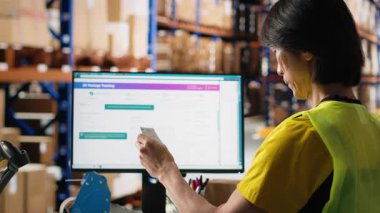 Asyalı bir çalışan, kargo paketlerinden önce havayolu fatura etiketlerinden takip bilgilerini daktilo ediyor, endüstriyel ambardaki sipariş tamamlama işlemini yönetiyor. B kameralı büyük iş deposu.