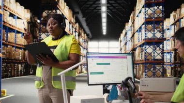 Siyahi müşteri hizmetleri çalışanı müşterilerle kargo sorunları hakkında konuşuyor, endüstriyel depodaki paket bilgilerine yardım ediyor. Kadın, sipariş karışıklığını kulaklıkla çözer. Kamera A.