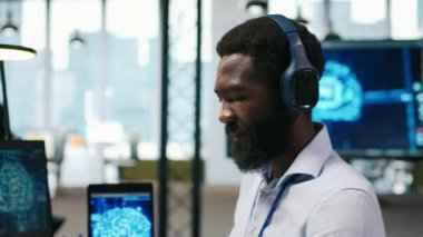 Ofisteki bilgisayar bilimcisi yapay zeka öğrenme sistemlerini iş akışlarını düzene sokacak şekilde entegre ediyor, kulaklık takıyor. Afrikalı Amerikalı adam cihazlar, dinleme müziği ve kamera A üzerinde yapay zeka kullanıyor.