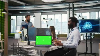 Ofis arkadaşlarının yanındaki teknisyen yeşil ekran laptopunu kullanarak yapay zeka otomasyonunu entegre ederek yazılım uygulamaları geliştiriyor. Bilgi İşlem uzmanı, A kamerası ve maket defteri ile hata ayıklama uygulamaları yapıyor.