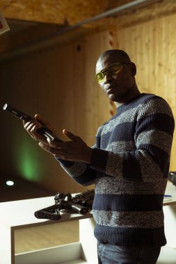 Poligon müşterisinin portresi. Tabanca şarjörünü dolduruyor. Hedefleri vurmaya hazırlanıyor, eğleniyor. Kapalı alanda ateş eden, tabancasını dolduran, koruyucu gözlüklü Afrikalı Amerikalı bir adam.