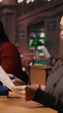 Ciddi bir ifadeye sahip Dikey Video Kadını, edebiyat anlayışını zenginleştirmek için akademik araştırmalar yapan ve ders kitapları okuyan bir halk kütüphanesi masasında makaleler okuyor. Kendi kendine çalışma. Kamera.