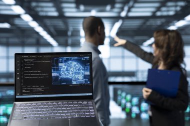 Veri merkezi teknisyenlerinin önündeki laptop bulanık arkaplandaki AI sürgülü yönetim araçları, sunucu kümelerini denetleme, ağ topolojilerini güncelleme, depolama bütünlüğünü doğrulama