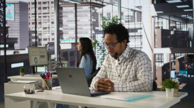 Siyahi bir çalışan ofis masasındaki bir telekomünikasyon toplantısına katılıyor, proje yönetimi ve iş stratejileri arasında birden fazla iş yapıyor. Bölümler arası verimlilik ve işbirliği. Kamera B.