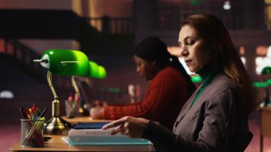 Kitaplar ve akademik materyallerle çevrili, hafif ışıklandırma altında okuyan yaşlı bir kadınla halk kütüphanesi ayarları. Üniversite mezunu olma yolunda kendini adamış bir kişi. Kamera B.