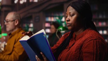 Afrikalı Amerikalı kadın kitap okuyor ve ders notlarını çalışma odasında tamamlıyor, tezine hazırlanıyor ve halk kütüphanesinde arşivlenmiş akademik materyalleri araştırıyor. Kamera B.